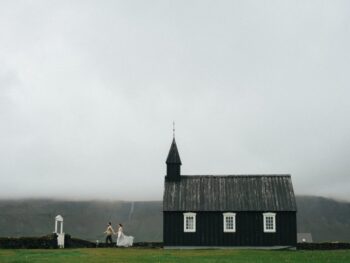 This tiny black church rising from a lava field is moody Iceland at its finest as a married couple walks out of the church. she is wearing a white dress and he is wearing a shirt and pants