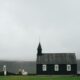 This tiny black church rising from a lava field is moody Iceland at its finest as a married couple walks out of the church. she is wearing a white dress and he is wearing a shirt and pants