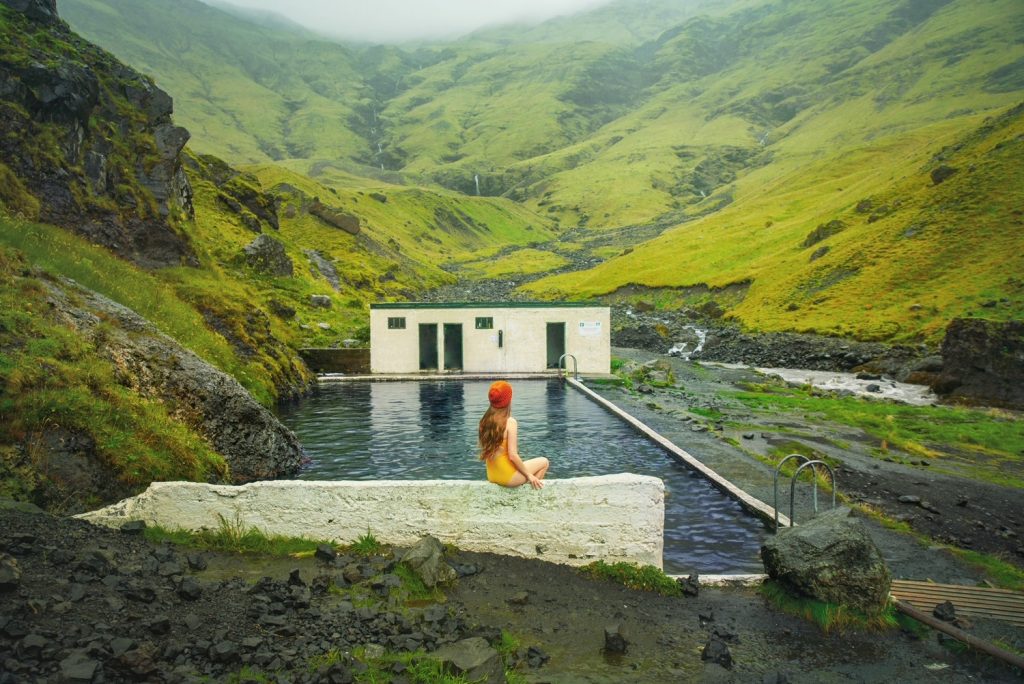 a girl in yellow swimsuit sitting on a concrete stone near a geothermal pool in the valley surrounded by rolling green hills