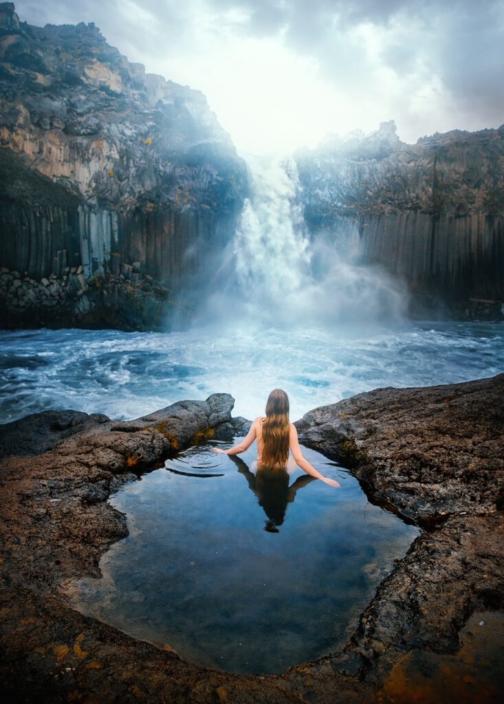 a girl with her back turned towards camera soaking in a hot spring with a waterfall in the background