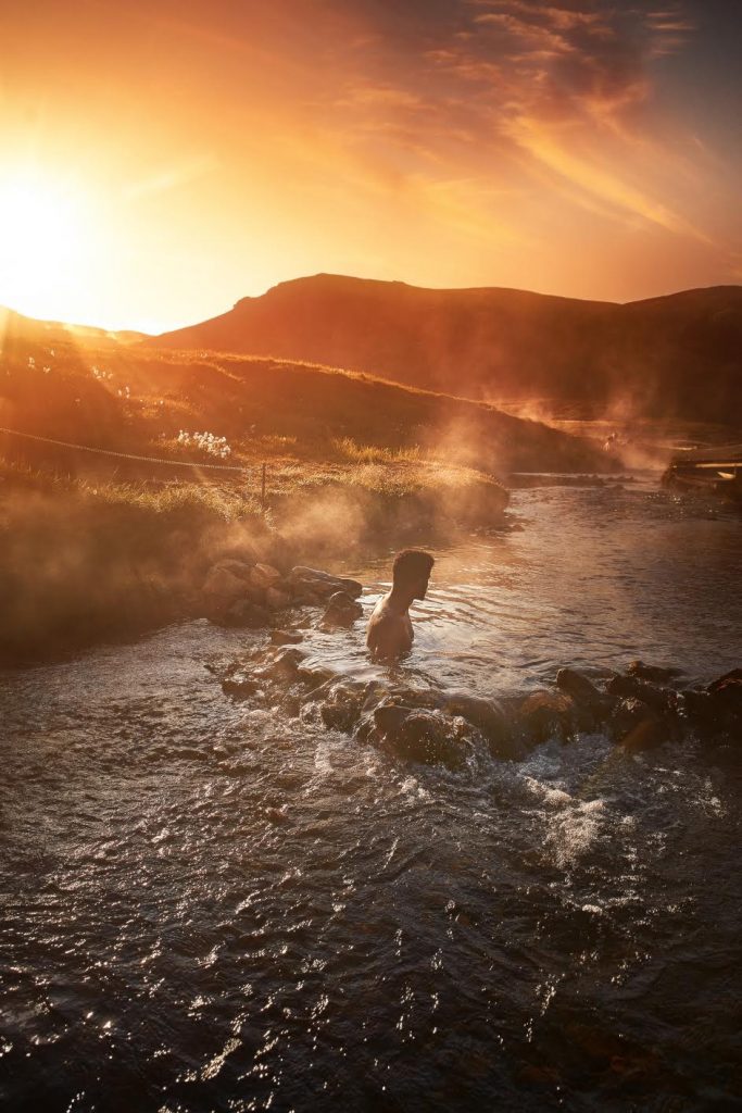 a man soaking in the hot spring at sunset with orange sky and mountains in the background