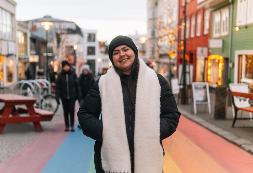 A woman smiles in her puffy jacket, scarf, and beanie as she is ready for what to wear in Reykjavik while standing on the rainbow road in cold weather. 