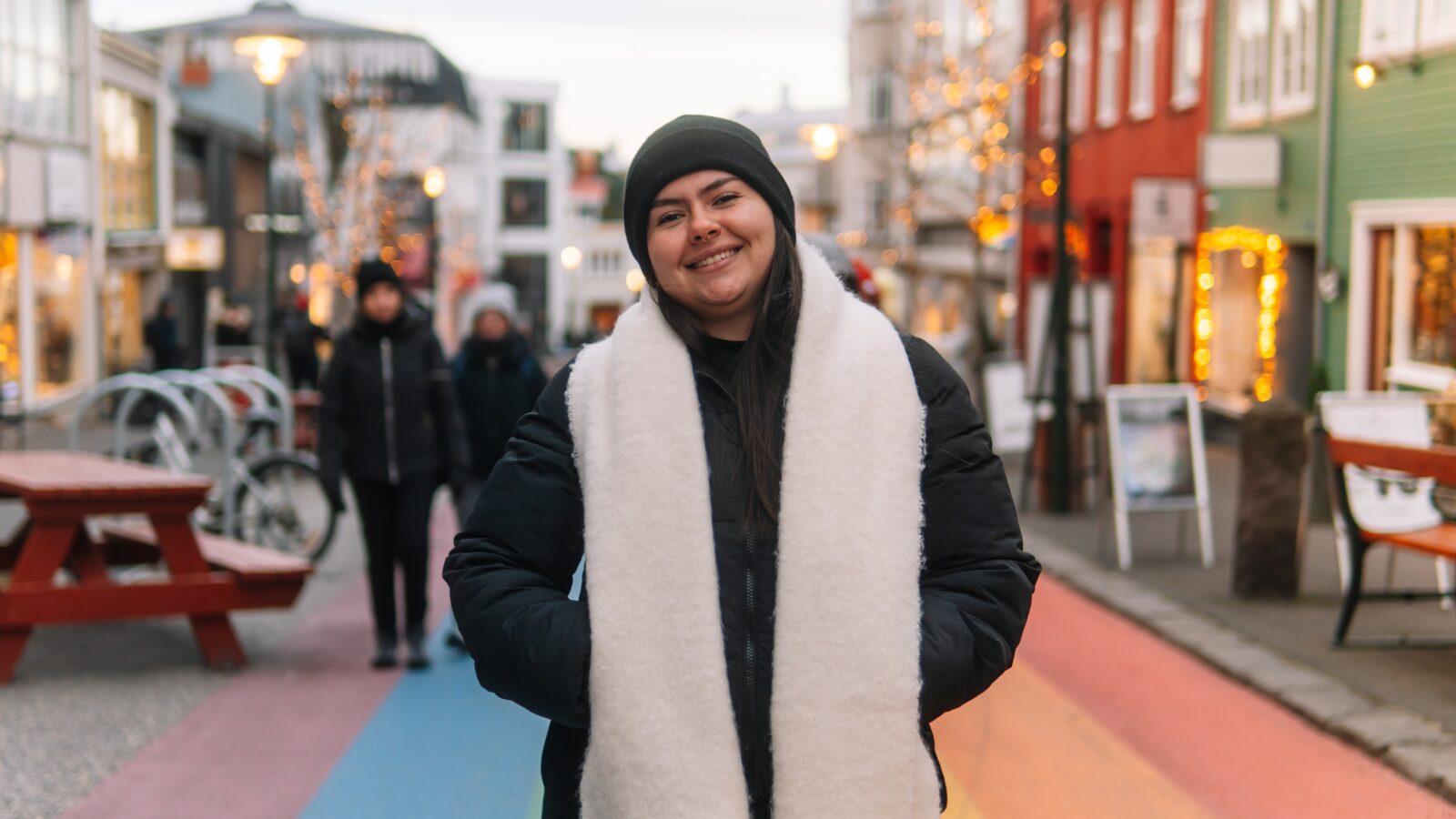 A woman knows what to wear in Reykjavik to stay warm: she has a beanie on, a scarf, and a puffer jacket as she stands on Reykjavik's famous rainbow road!