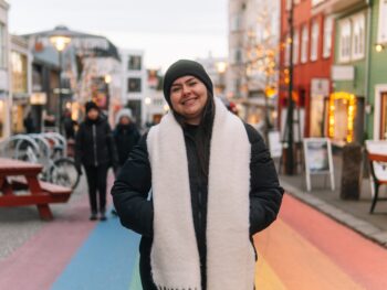 A woman knows what to wear in Reykjavik to stay warm: she has a beanie on, a scarf, and a puffer jacket as she stands on Reykjavik's famous rainbow road!