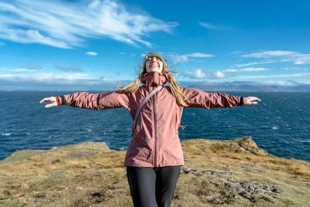 A woman spreads her arms out and enjoys the sun and warmth on the coast, but still knows what to wear in Reykjavik, with her zip up windbreaker and beanie! 