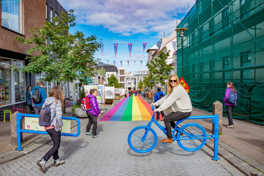 A woman in a sweater and insulated pants knows what to wear in Reykjavik even. in summer, as she poses on a bike statue by the rainbow road in Reykjavik. 