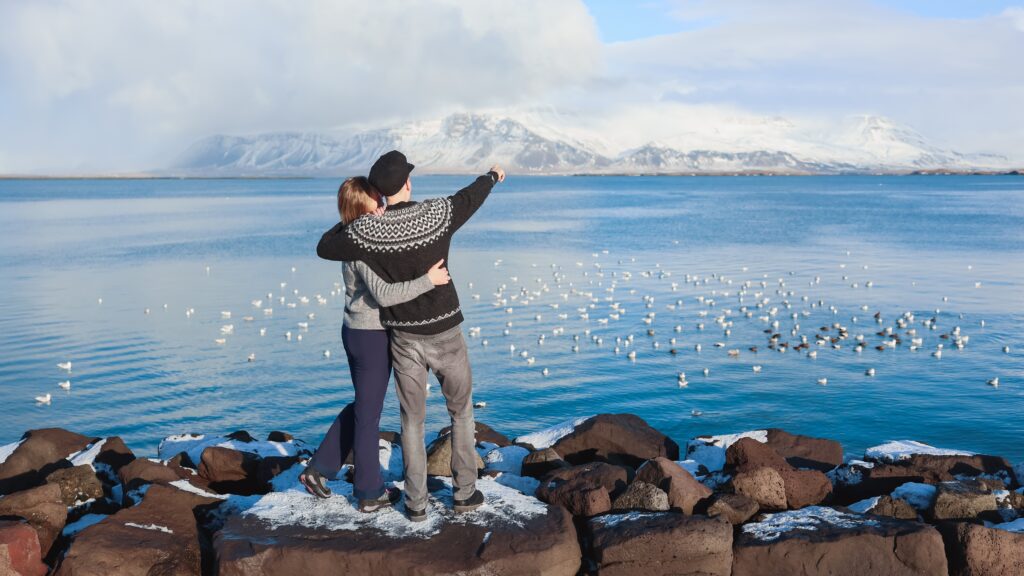 A couple in hiking pants and classic Icelandic sweaters stand on a rocky terrain and point across the water, prepared in what to wear in Reykjavik. 