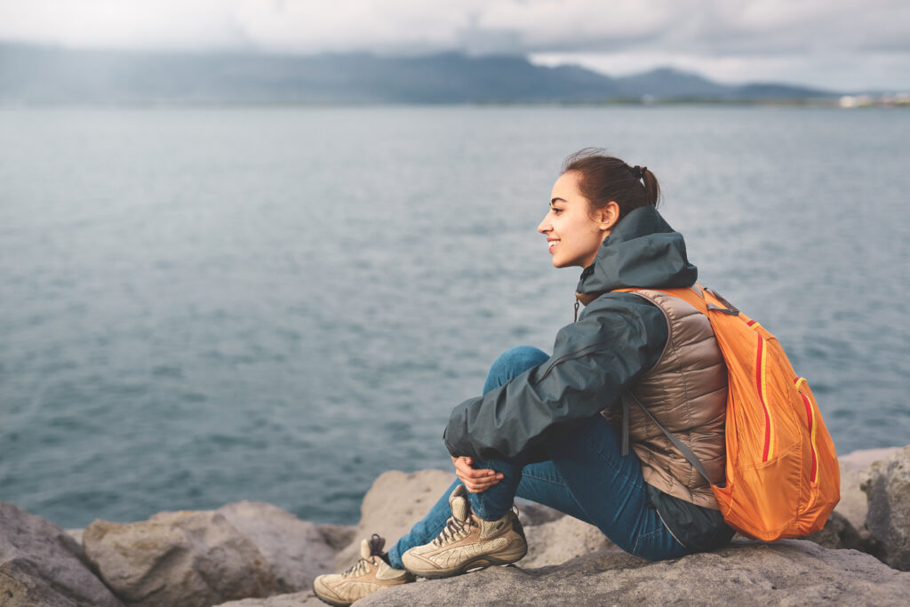 A woman sits on a rick and overlooks water on a foggy day in a vest, waterproof jacket, hiking boots and orange backpack. 